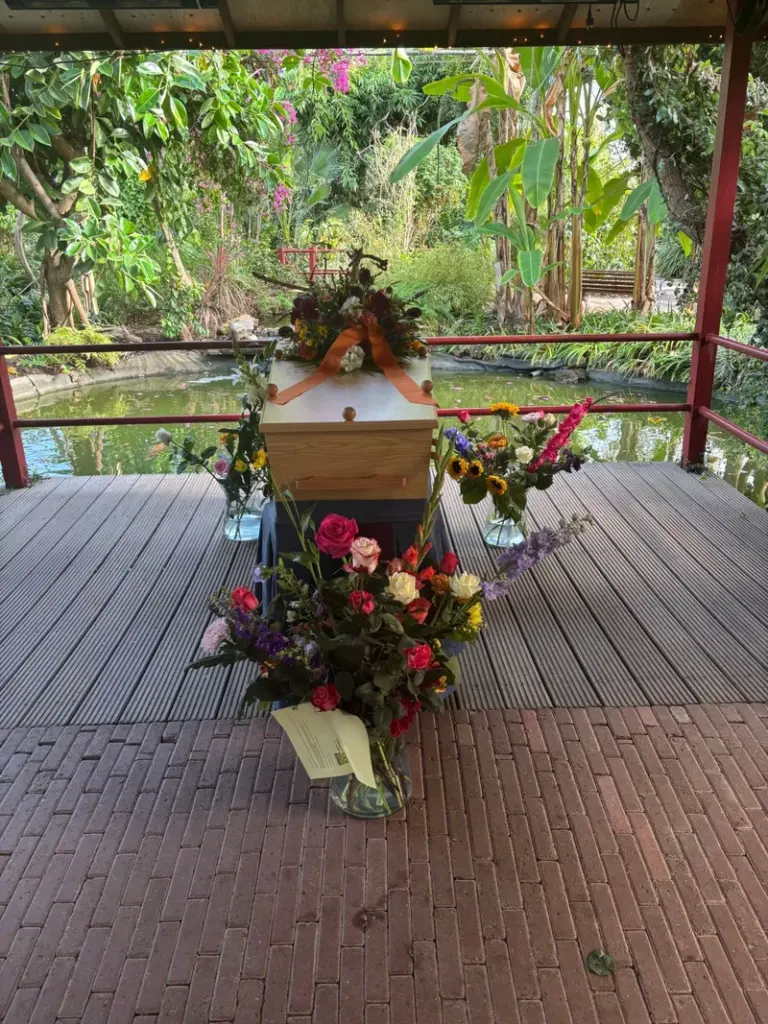 Een lichte kist met een bloemstuk en een oranje lint op een vlonder aan een vijver, onder een overkapping tussen tropische planten, met vazen vol bloemen eromheen