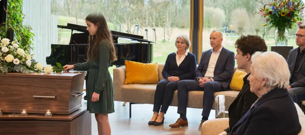 Een meisje legt een witte roos op een kist in een lichte zaal met grote ramen, familie kijkt toe vanaf een bank