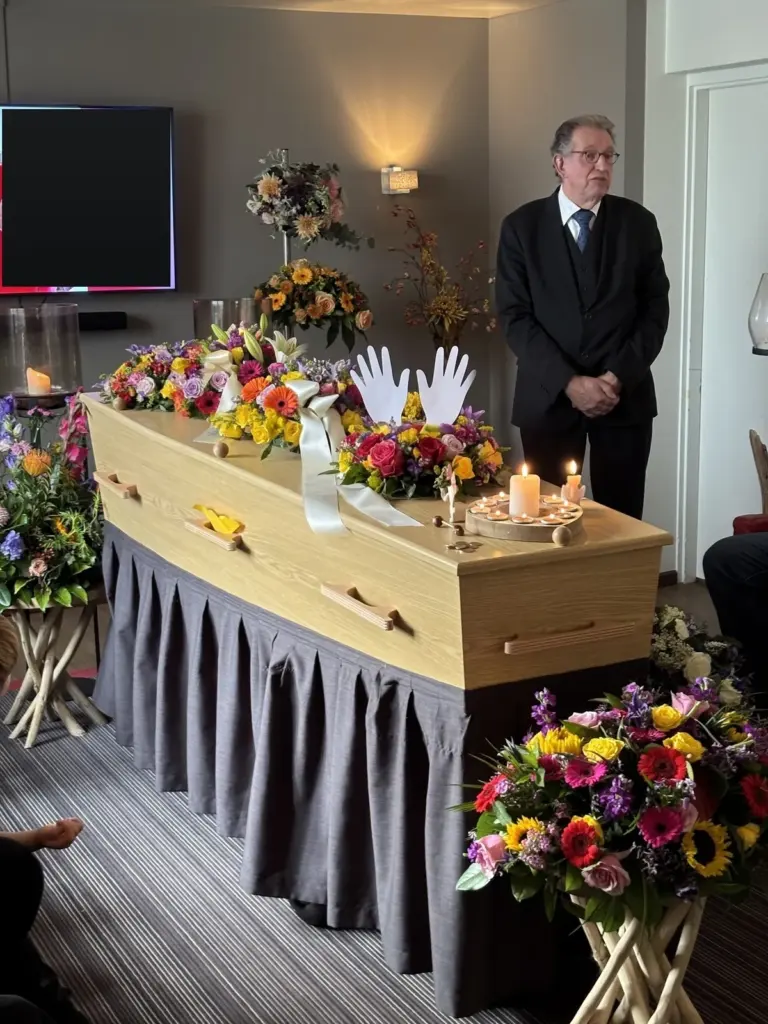 Martin Uitendaal staat naast een kist vol bloemen in een afscheidsruimte, met een brandende kaars
