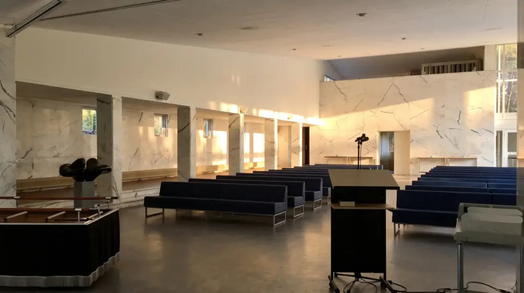 De aula van begraafplaats Duinhof met marmeren wanden, rijen donkerblauwe banken en een lessenaar in ochtendlicht