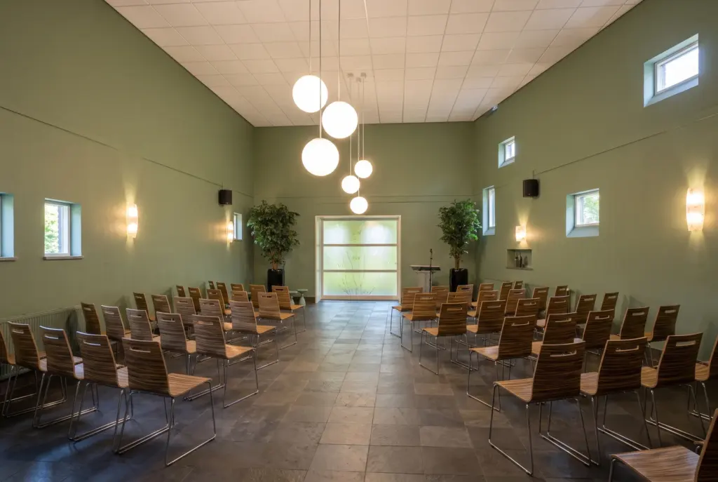 De aula van begraafplaats Houtrijk en Polanen met groene wanden, witte bollampen en rijen houten stoelen aan weerszijden van een middenpad