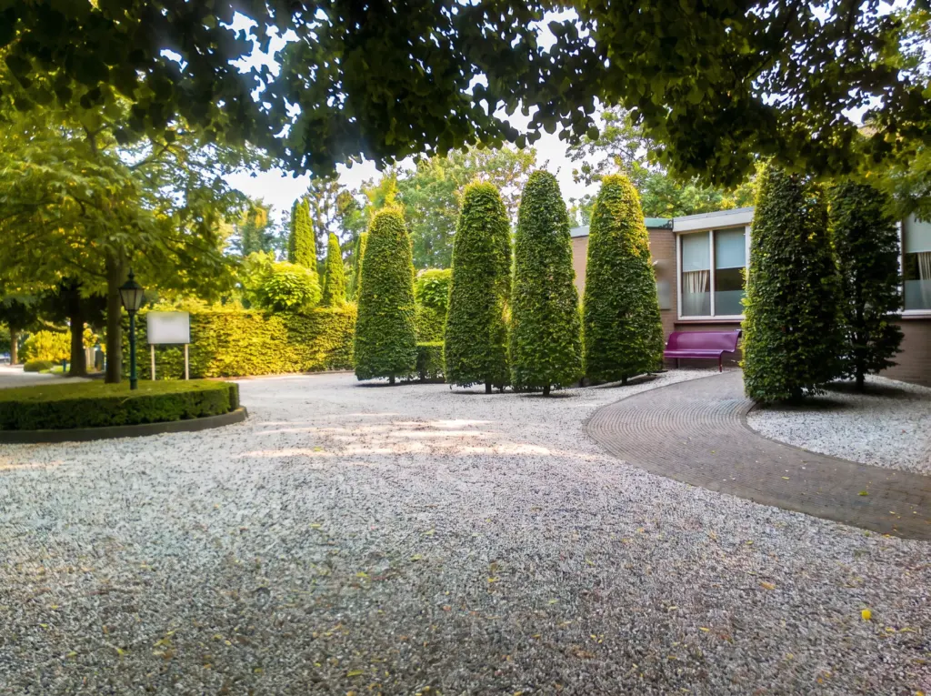 Het voorplein van begraafplaats Houtrijk en Polanen in Zwanenburg: grind, zuilvormig geschoren haagbeuken en het lage bakstenen aulagebouw