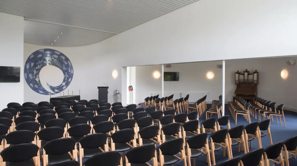De aula van begraafplaats Noorderduin met rijen stoelen, een blauw rond mozaïek aan de wand en een houten orgel