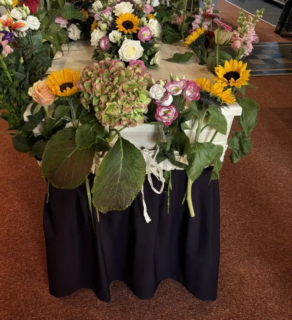 Een kist vol zonnebloemen, rozen en hortensia’s op een baar met een donkerblauwe rok, in een kerk