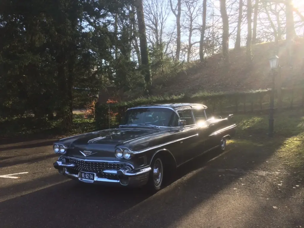 De donkergrijze Cadillac Fleetwood uit 1958 van Uitendaal, met vier koplampen en staartvinnen, in de lage ochtendzon tussen de bomen