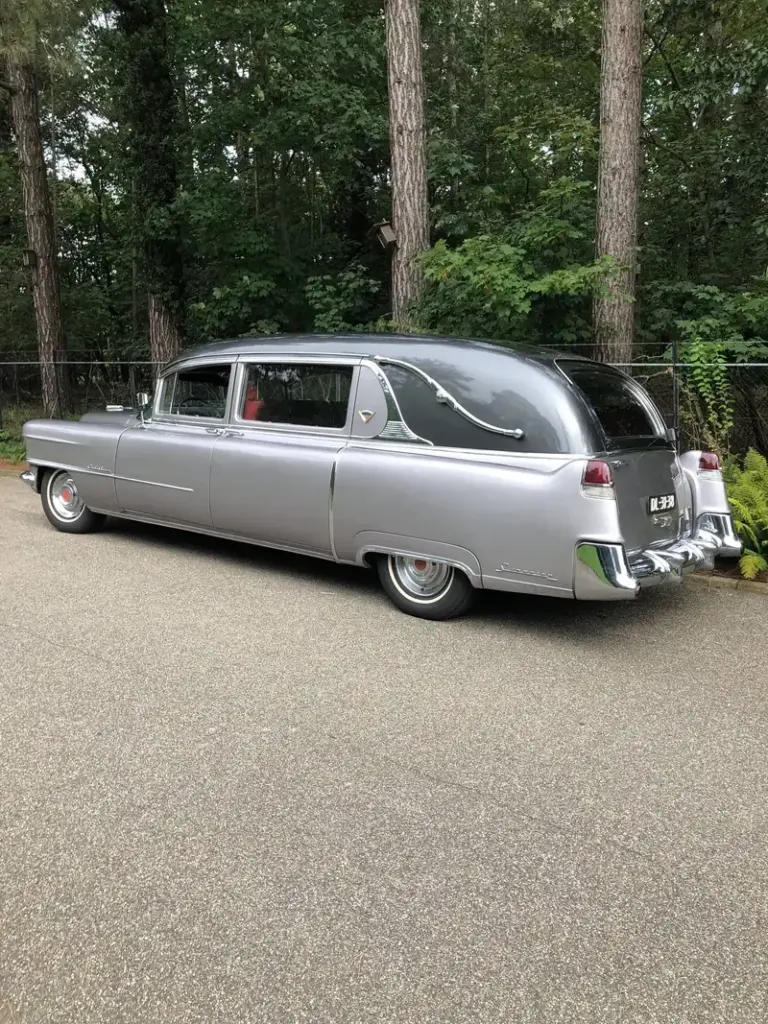 Een zilvergrijze Cadillac rouwauto uit 1955 van opzij, geparkeerd langs een bosrand
