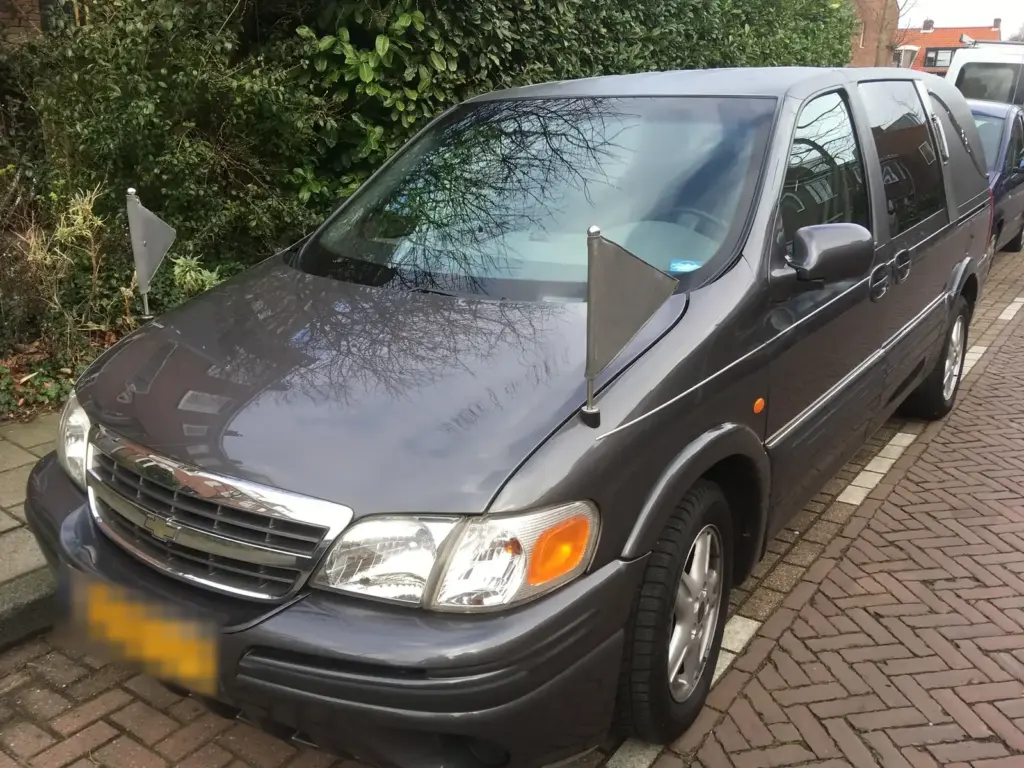Een grijze Chevrolet uit het wagenpark van Uitendaal met twee rouwvlaggetjes op de motorkap, geparkeerd in een straat met klinkers