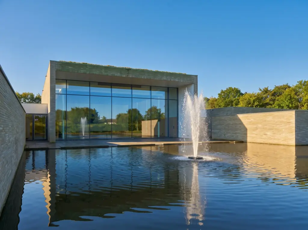 Crematorium Duin en Bollenstreek in Lisse: een laag stenen gebouw met een glazen gevel aan een vijver met fontein, onder een blauwe lucht