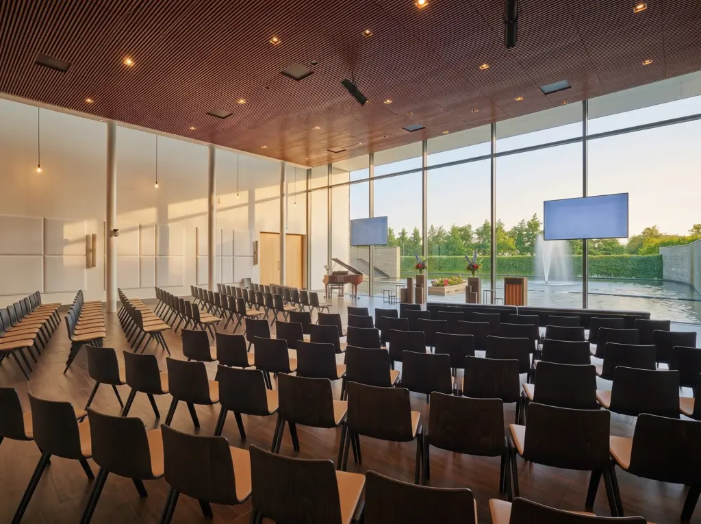 De Rembrandtzaal van Crematorium Duin en Bollenstreek met rijen stoelen en een glazen wand naar de vijver