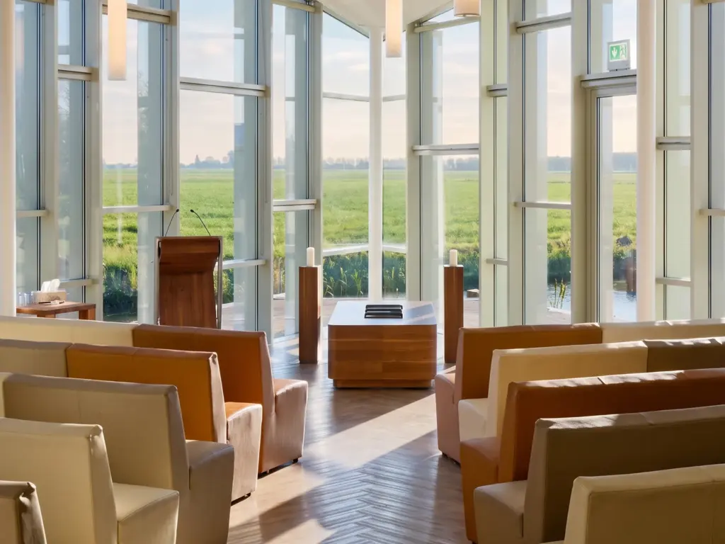 De grote aula gezien vanaf de achterste rijen: fauteuils in crème en cognac, een houten sokkel voor de kist en een glazen wand met uitzicht over de polder