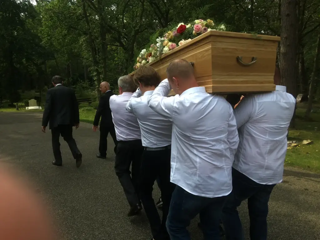 Dragers in witte overhemden dragen een kist met bloemen over een pad op een begraafplaats