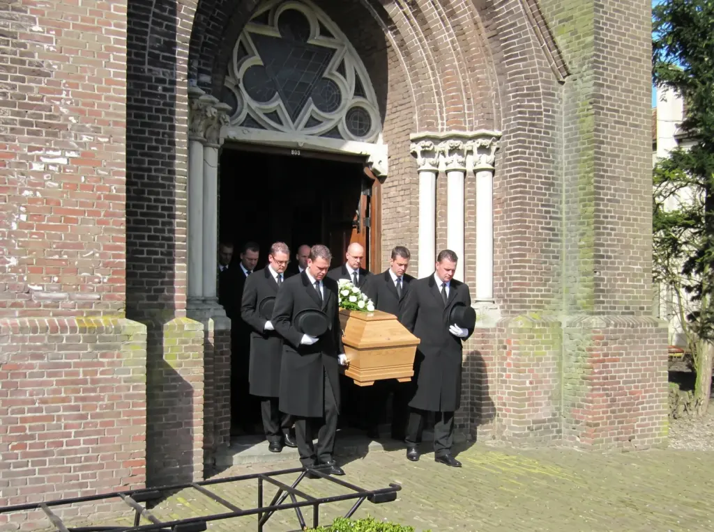 Dragers in lange zwarte jassen, met de hoge hoed in de hand, dragen een eikenhouten kist door de poort van een bakstenen kerk naar buiten