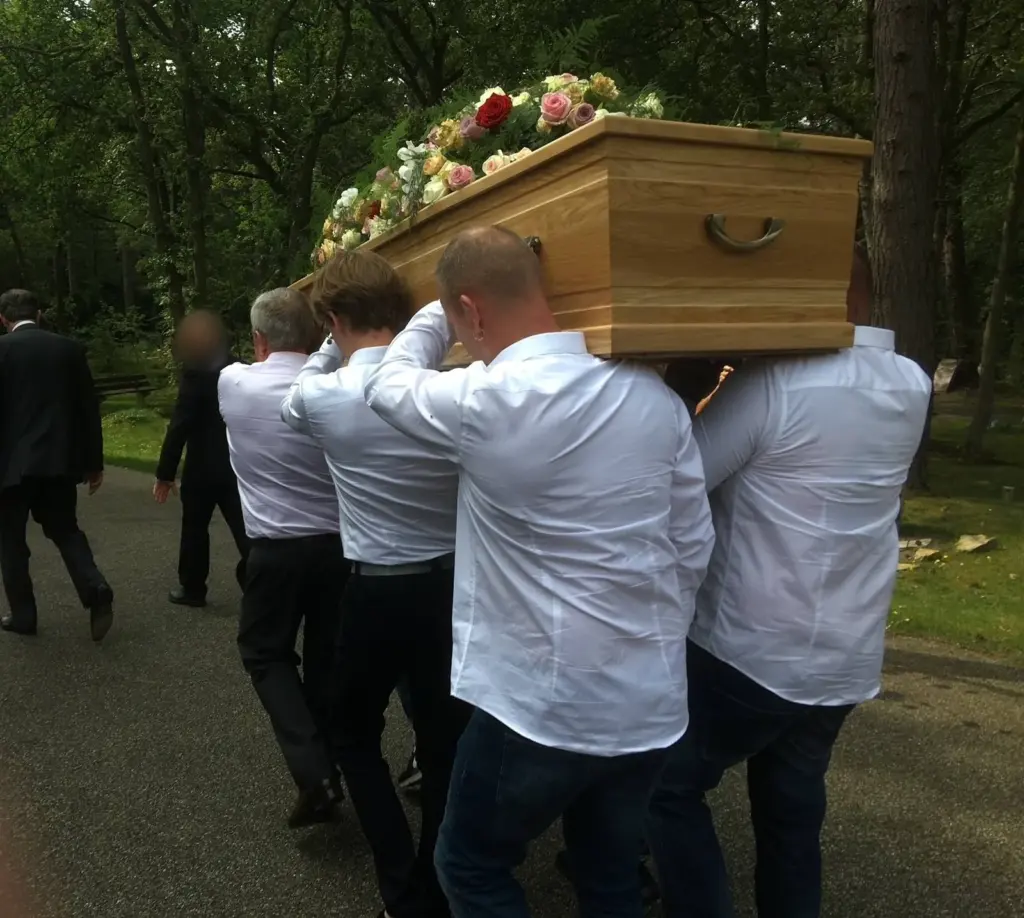 Dragers in witte overhemden brengen een eikenhouten kist met bloemen over het pad van de begraafplaats