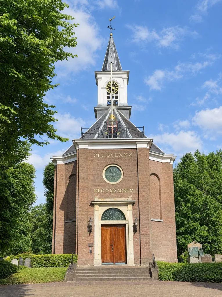 De Hervormde Kerk van Bennebroek: een bakstenen kerk met witte lijsten, een wit klokkentorentje met windvaan, het jaartal in Romeinse cijfers en een houten deur boven een stenen trap, tussen hoge bomen