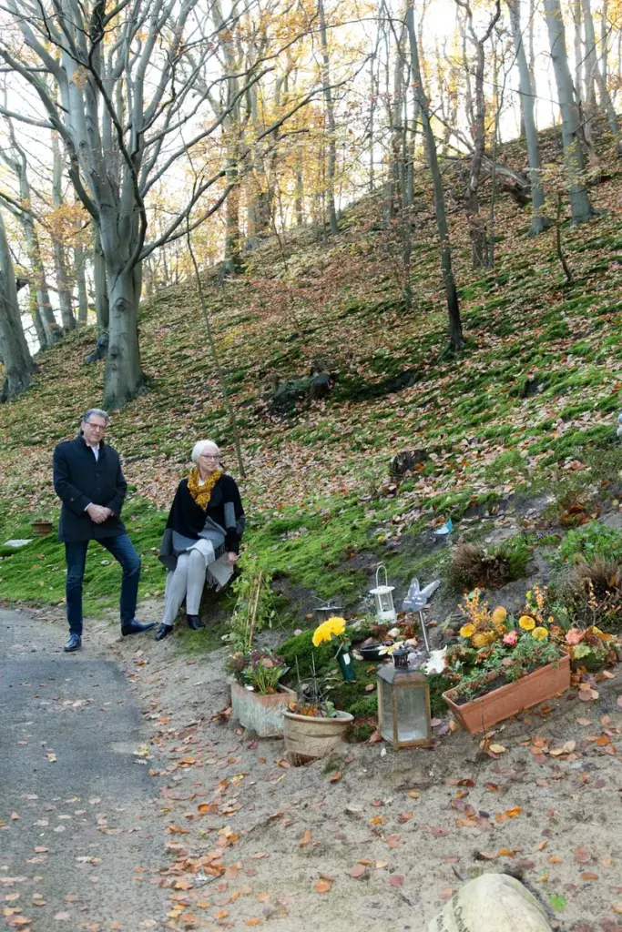 Martin Uitendaal en Sonja Raijmakers op een beboste helling naast een graf met bloemen en lantaarns