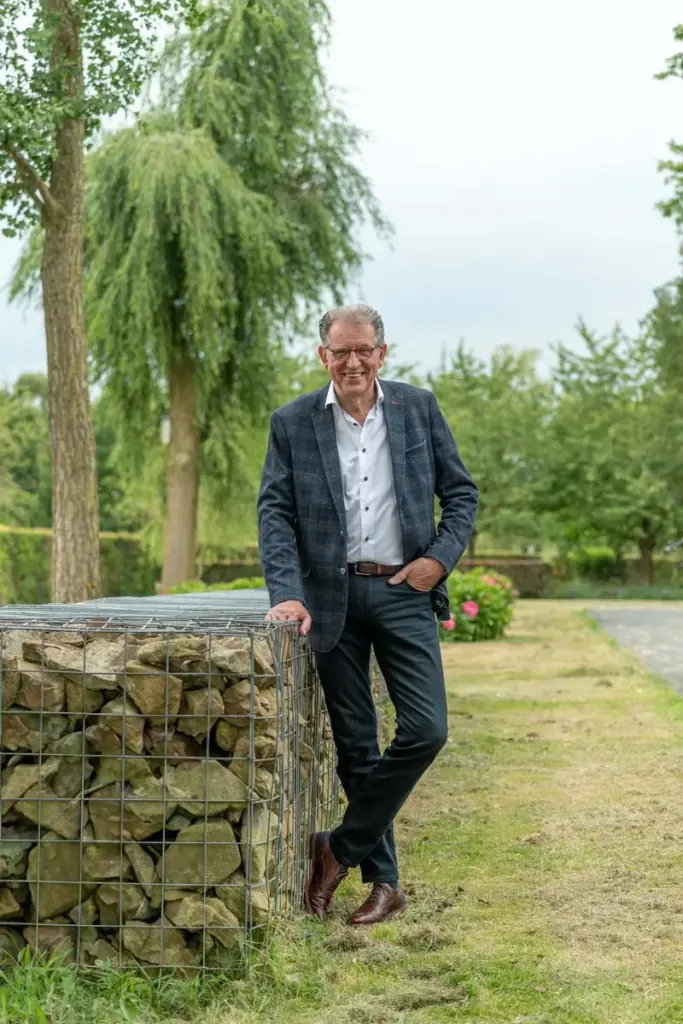 Martin Uitendaal leunt tegen een stenen muurtje in een tuin met een treurwilg