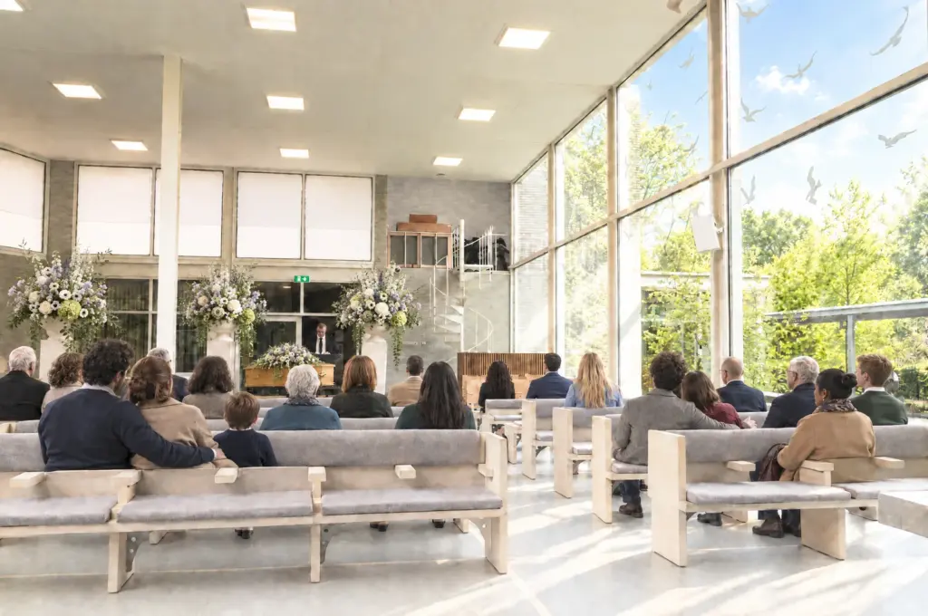 Familie en vrienden van jong tot oud, in ingetogen kleding, op de lichte houten banken van de aula van Rietveld, met voorin de kist en de spreker tussen bloemstukken in wit en lila, en rechts de hoge glazen wand met witte vogelsilhouetten