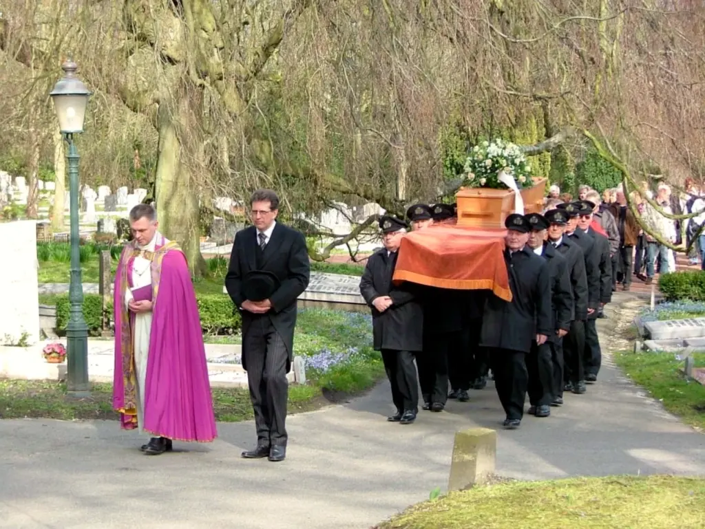 Een oudkatholieke begrafenis: een geestelijke in paarse koorkap en Martin Uitendaal met de bolhoed in de hand lopen voor de dragers in zwarte jassen en uniformpetten, die de kist met een oranje kleed dragen, gevolgd door familie en belangstellenden