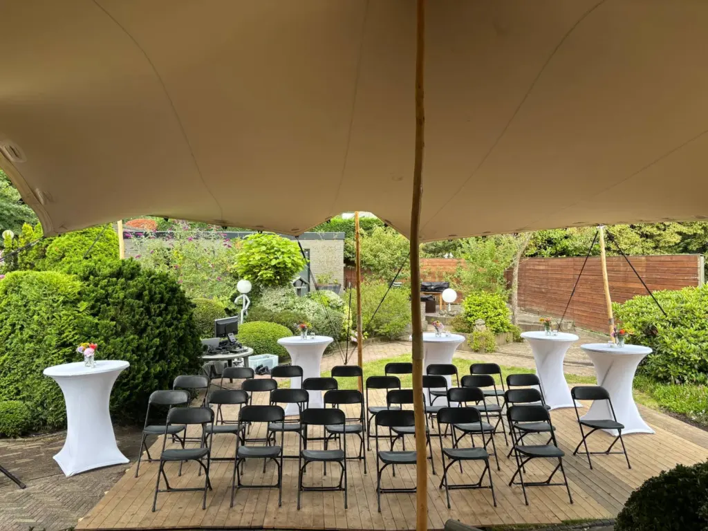 Rijen zwarte klapstoelen en statafels met witte hoezen en kleine boeketjes onder een tentdoek in een tuin, klaar voor een plechtigheid