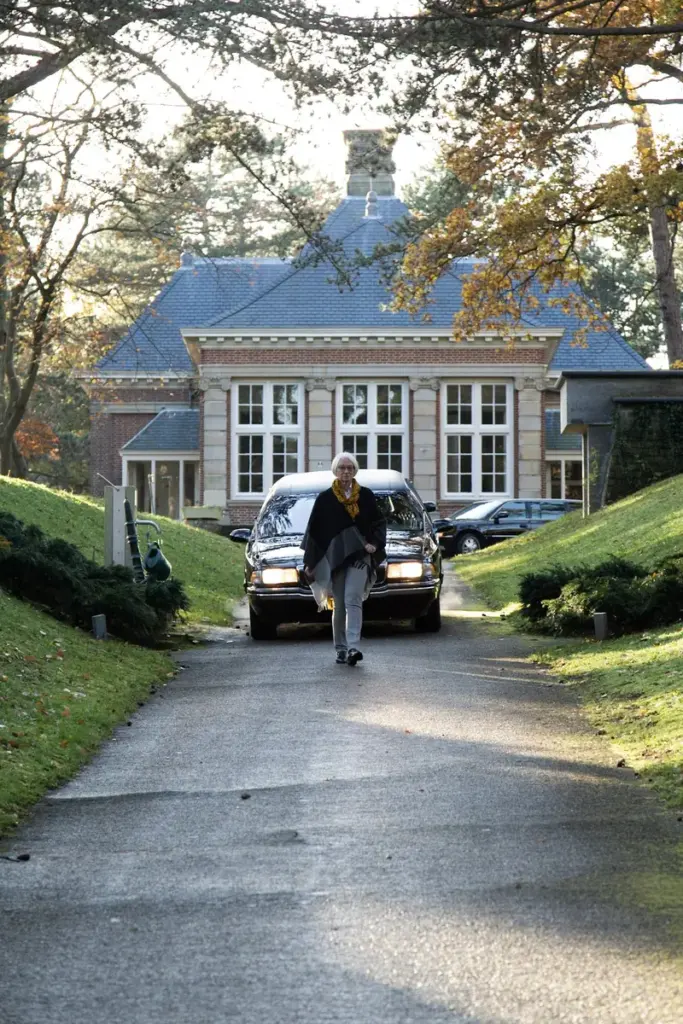 Een rouwauto staat op de oprijlaan voor een aula tussen hoge bomen