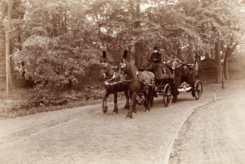 Sepiafoto van een zwarte rouwkoets met lantaarns en gedrapeerde gordijnen, getrokken door twee Friese paarden met pluimen, met een koetsier in hoge hoed op een klinkerpad door het bos